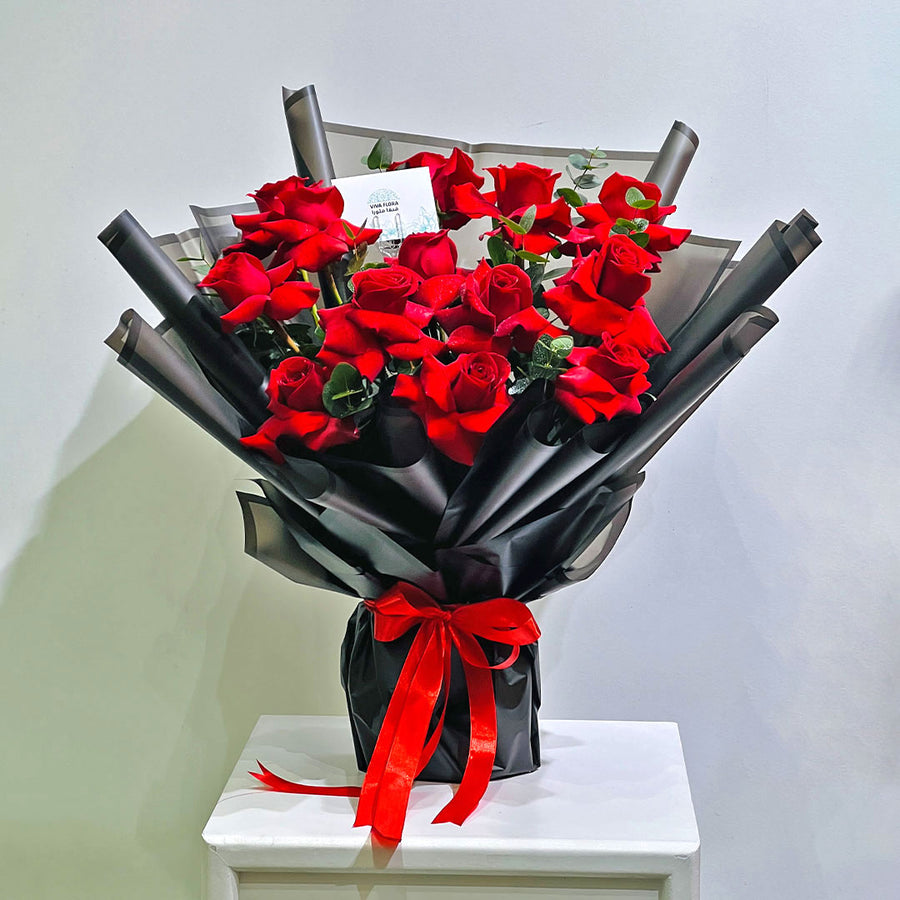 A large bouquet of red roses wrapped in black paper and tied with a red ribbon, displayed on a white table against a plain light gray wall.