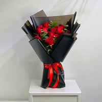 A bouquet of red roses wrapped in black paper with a bright red ribbon, displayed on a white pedestal against a light gray wall.