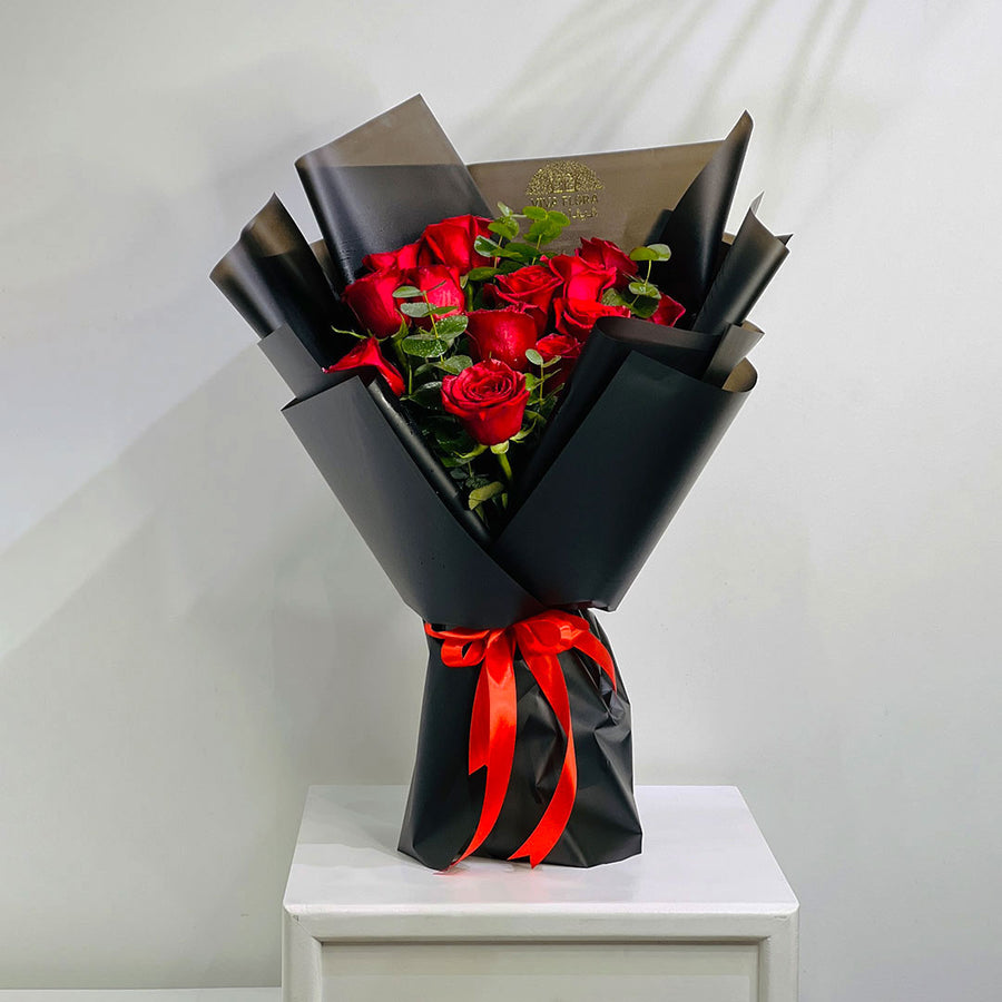 A bouquet of red roses wrapped in black paper with a bright red ribbon, displayed on a white pedestal against a light gray wall.