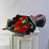 A bouquet of 15 red roses wrapped in black paper with a red ribbon, displayed on a white cabinet against a light gray wall.
