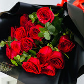 A bouquet of red roses wrapped in black paper with a small gold floral label, photographed close up against a light neutral background.