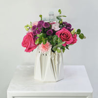 A floral arrangement in a white paper bag-style vase, featuring pink and purple flowers with green foliage, displayed on a white surface against a light gray background.