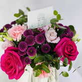 A vibrant floral bouquet featuring bright pink and magenta roses, light pink carnations, deep purple chrysanthemums, and green eucalyptus leaves, arranged in a white vase with a 'VIVA FLORA' card.