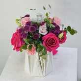 A vibrant floral bouquet featuring bright pink and magenta roses, light pink carnations, deep purple pompons, and green eucalyptus leaves, arranged in a white geometric paper vase with a brand card insert.