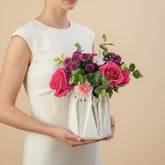 A floral arrangement in a white paper bag-style vase, featuring pink and purple flowers with green foliage, displayed on a white surface against a light gray background.