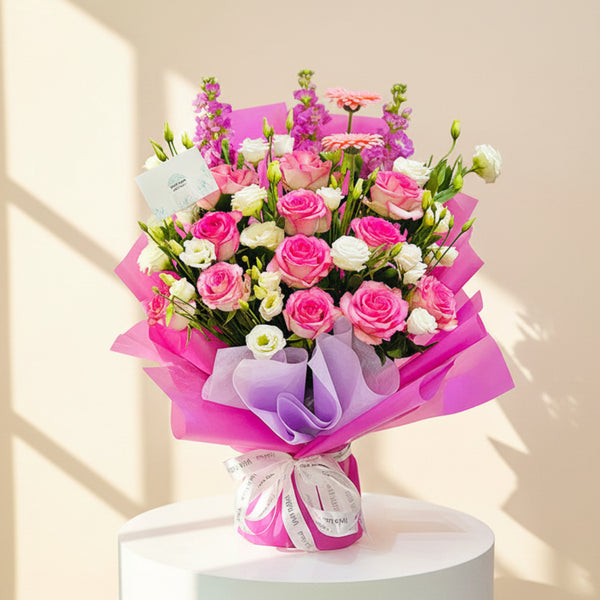 A vibrant floral bouquet wrapped in bright pink and purple tissue paper, featuring pink and white roses, pink gerbera daisies, and purple delphiniums, displayed on a white pedestal against a light gray background.