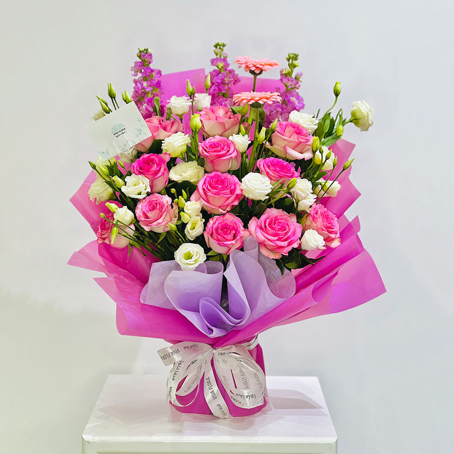 A vibrant floral bouquet wrapped in bright pink and purple tissue paper, featuring pink and white roses, pink gerbera daisies, and purple delphiniums, displayed on a white pedestal against a light gray background.