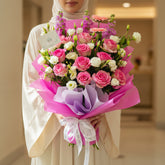 A vibrant floral bouquet wrapped in bright pink and purple tissue paper, featuring pink and white roses, pink gerbera daisies, and purple delphiniums, displayed on a white pedestal against a light gray background.