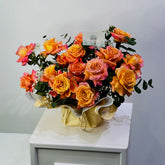A vibrant bouquet of orange and pink roses with green foliage, wrapped in gold paper, displayed on a white cabinet against a light gray wall.