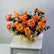A vibrant bouquet of orange and pink roses with green foliage, wrapped in gold paper, displayed on a white cabinet against a light gray wall.