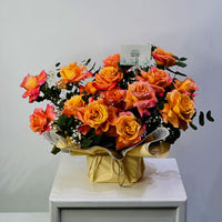 A large floral arrangement of orange, pink, and yellow roses with green foliage, wrapped in gold paper and tied with a bow, displayed on a white cabinet against a light gray wall.