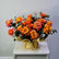 A large floral arrangement of orange, pink, and yellow roses with green foliage, wrapped in gold paper and tied with a bow, displayed on a white cabinet against a light gray wall.