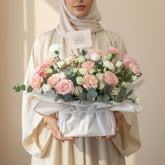 A floral arrangement of pink and white roses, carnations, and green foliage in a white wrapped basket, displayed on a white pedestal against a warm beige wall with soft window shadows.