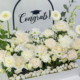 A graduation floral arrangement featuring cream and white roses, daisies, and carnations with green foliage, set in a white planter with small white stones. A black arch backdrop displays the word 'Congrats!' with a graduation cap graphic.