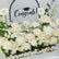 A graduation floral arrangement featuring cream and white roses, daisies, and carnations with green foliage, set in a white planter with small white stones. A black arch backdrop displays the word 'Congrats!' with a graduation cap graphic.
