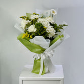 A bouquet of white roses and green foliage wrapped in white tissue paper and light green paper with a white ribbon, displayed on a white pedestal against a light gray background.