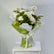A bouquet of white roses and green foliage wrapped in white tissue paper and light green paper with a white ribbon, displayed on a white pedestal against a light gray background.