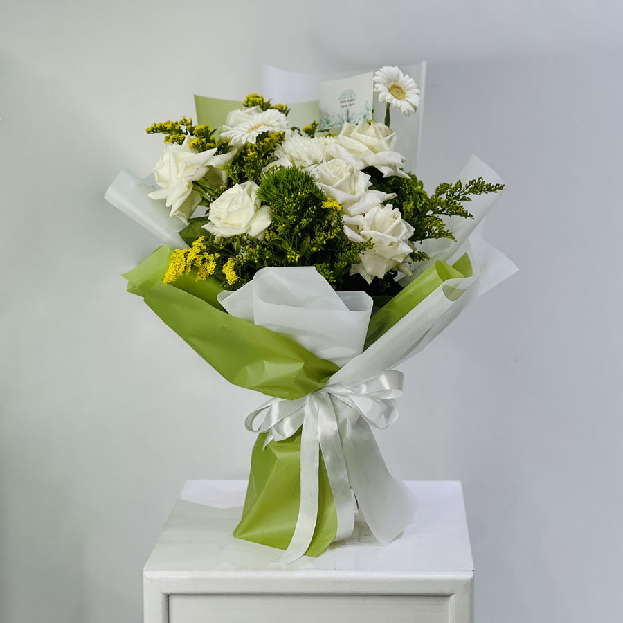 A bouquet of white roses and green foliage wrapped in white tissue paper and light green paper with a white ribbon, displayed on a white pedestal against a light gray background.