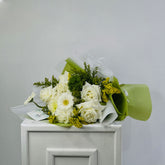 A bouquet of white flowers, including roses and daisies, wrapped in white tissue paper and light green paper, resting on top of a white cabinet against a plain light gray wall.