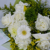 A close-up bouquet of white roses, a white daisy with a yellow center, green filler flowers, and small yellow blossoms, with part of a greeting card visible at the lower left.