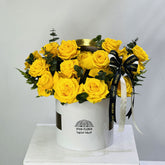 A bouquet of bright yellow roses arranged in a white cylindrical box with a black and white ribbon, displayed on a white pedestal against a light gray background. The box is branded VIVA FLORA.