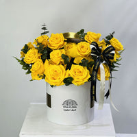 A bouquet of bright yellow roses arranged in a white cylindrical box with a black and white ribbon, displayed on a white pedestal against a light gray background. The box is branded VIVA FLORA.