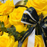 A close-up of a yellow rose bouquet with green eucalyptus leaves and a black ribbon printed with the brand name VIVA FLORA.