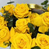 A close-up arrangement of bright yellow roses with green eucalyptus stems in a dark container with a gold rim, photographed against a light neutral background.