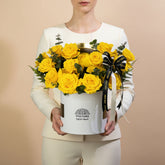 A bouquet of bright yellow roses arranged in a white cylindrical box with a black and white ribbon, displayed on a white pedestal against a light gray background. The box is branded VIVA FLORA.