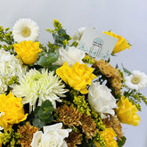 A vibrant floral arrangement featuring bright yellow roses, white daisies, white roses, and white chrysanthemums, accented by green eucalyptus leaves and small brown chrysanthemums, with a small white card in the center.
