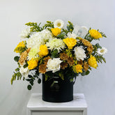 A vibrant floral arrangement featuring yellow roses, white daisies, white carnations, and orange-brown chrysanthemums in a black cylindrical box, displayed on a white pedestal against a light gray background.