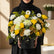 A vibrant floral arrangement featuring yellow roses, white daisies, white carnations, and orange-brown chrysanthemums in a black cylindrical box, displayed on a white pedestal against a light gray background.