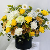 A vibrant floral arrangement featuring yellow roses, white daisies, white roses, and white chrysanthemums, styled in a black cylindrical box on a white marble surface against a light gray background.