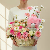 A wicker basket floral arrangement with pink and white flowers, a pink teddy bear, and a silver glittery plush toy, displayed on a white pedestal against a beige wall with window shadow light.