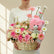 A wicker basket floral arrangement with pink and white flowers, a pink teddy bear, and a silver glittery plush toy, displayed on a white pedestal against a beige wall with window shadow light.