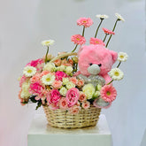 A floral gift basket arrangement featuring a pink teddy bear plush toy, pink and white daisies, pink and white roses, and a wicker basket on a white pedestal against a light gray background.