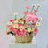A floral gift basket arrangement featuring a pink teddy bear plush toy, pink and white daisies, pink and white roses, and a wicker basket on a white pedestal against a light gray background.