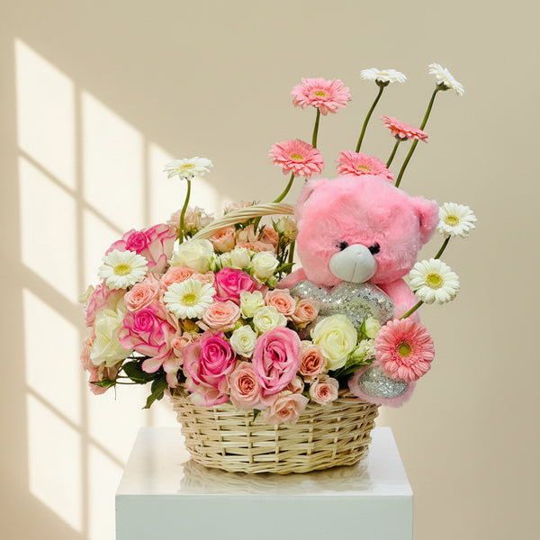 A floral gift basket arrangement featuring a pink teddy bear plush toy, pink and white daisies, pink and white roses, and a wicker basket on a white pedestal against a light gray background.