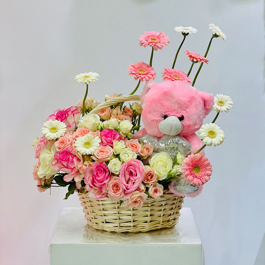 A floral gift basket arrangement featuring a pink teddy bear plush toy, pink and white daisies, pink and white roses, and a wicker basket on a white pedestal against a light gray background.