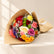 A colorful flower bouquet wrapped in brown kraft paper, featuring red, pink, yellow, orange, purple, and white flowers, placed on a white surface against a white marble-patterned wall.