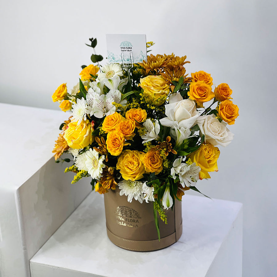 A floral arrangement of yellow and white roses, chrysanthemums, and green foliage in a round brown box, displayed on a white pedestal against a light gray background.