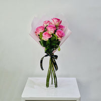 A bouquet of pink roses wrapped in pale pink paper with a black ribbon printed with gold text, displayed on a white pedestal against a light gray background.