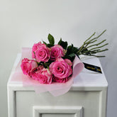 A bouquet of pink roses wrapped in pale pink tissue paper and a black ribbon, displayed on top of a white cabinet against a light gray wall.