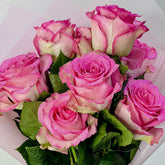 A bouquet of pink roses with green leaves, wrapped in white paper, shown in a close-up shot.