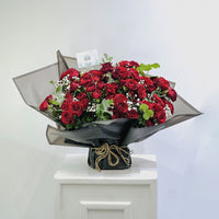 A bouquet of deep red roses with green leaves and small white baby's breath flowers, arranged in a gift box with a card that reads 'VIVA FLORA Arabic Love'.