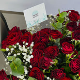 A bouquet of deep red roses with green leaves and small white baby's breath flowers, arranged in a gift box with a card that reads 'VIVA FLORA Arabic Love'.