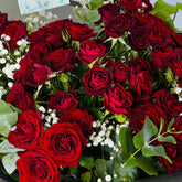 A dense bouquet of deep red roses with green leaves and small white baby's breath flowers, photographed close up.