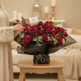 A large bouquet of red roses wrapped in black and gray paper with a small card tucked inside, displayed on a white pedestal against a plain light background.