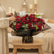 A large bouquet of red roses wrapped in black and gray paper with a small card tucked inside, displayed on a white pedestal against a plain light background.