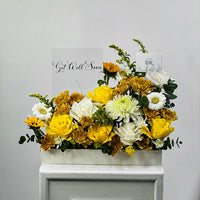 A rectangular white floral arrangement box filled with yellow and white flowers, including roses, chrysanthemums, and daisies, with green foliage. Two greeting cards are propped behind the arrangement against a light gray wall.